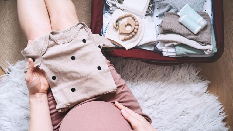 A pregnant person sitting on the floor, holding up a beige baby onesie with black polka dots while packing a hospital bag filled with baby clothes and essentials.