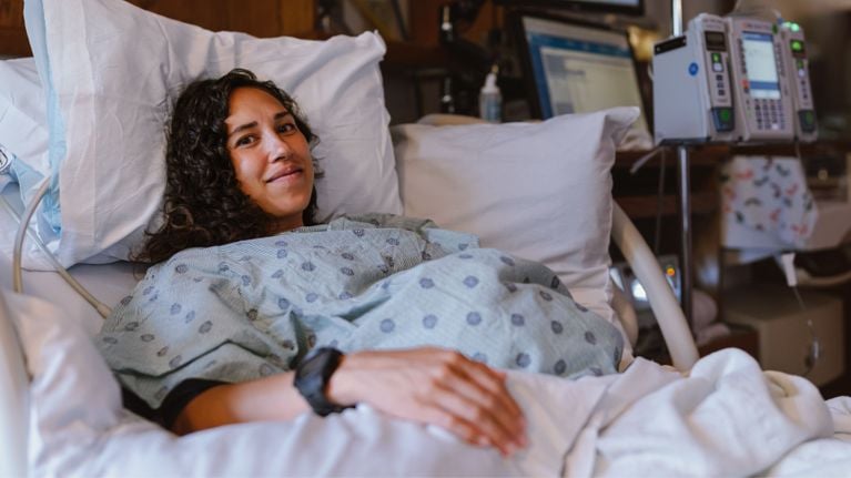 A pregnant person lies in a hospital bed wearing a gown, looking calm and smiling slightly, with medical monitors visible in the background.