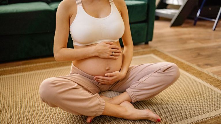 Pregnant person sitting cross-legged on a rug, holding their bare belly with both hands while wearing a sports bra and loose pants.