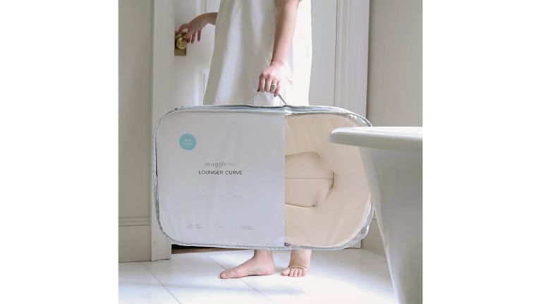 A person in a white dress carries a Snuggle Me Lounger Curve in a zip-up travel case, walking barefoot near a bathroom door.