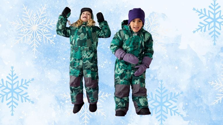 Two young children wearing green MEC Toaster Suits with a leafy print, standing and smiling in front of a winter-themed background. One child is jumping with joy, while the other poses playfully.