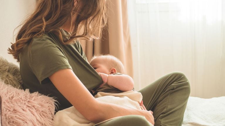 A woman in an olive green outfit breastfeeds her baby in a softly lit room. The baby is wrapped in a light blanket and nursing in a relaxed seated position.