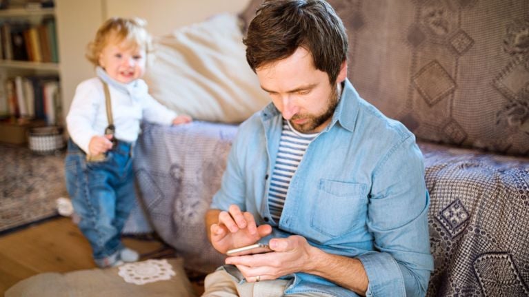 A father using his smartphone while sitting on the couch, with his toddler standing nearby looking at him.