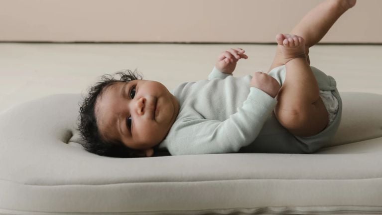 A baby wearing a white onesie and pink bow headband lies in a sage green baby lounger while a hand gently rests near their feet.