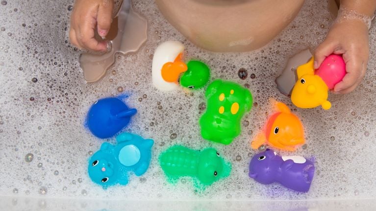 Close-up of a child’s hands holding rubber bath toys while sitting in a tub filled with foam and colorful floating animals.