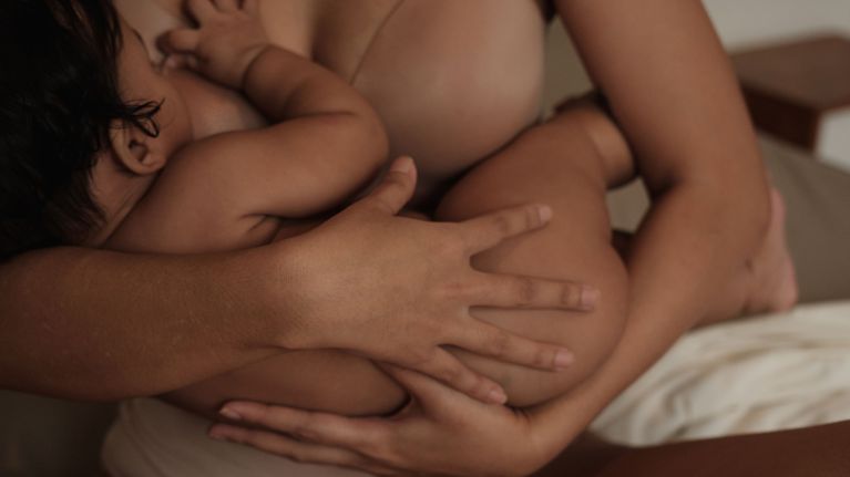 A close-up of a mother nursing her baby. The baby is latched on while the mother’s arms gently support and cradle the child.