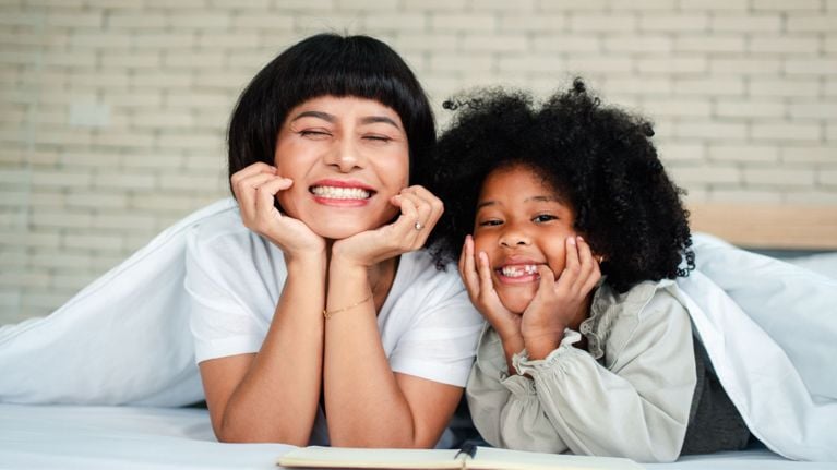 A mother and daughter lying on a bed, smiling brightly with their chins resting in their hands, enjoying time together.
