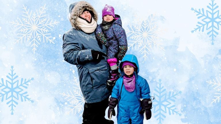 A parent bundled in a large blue winter coat stands in the snow with two children—one in a purple polka dot MEC suit and the other in a bright blue one-piece—with snow-covered trees in the background.