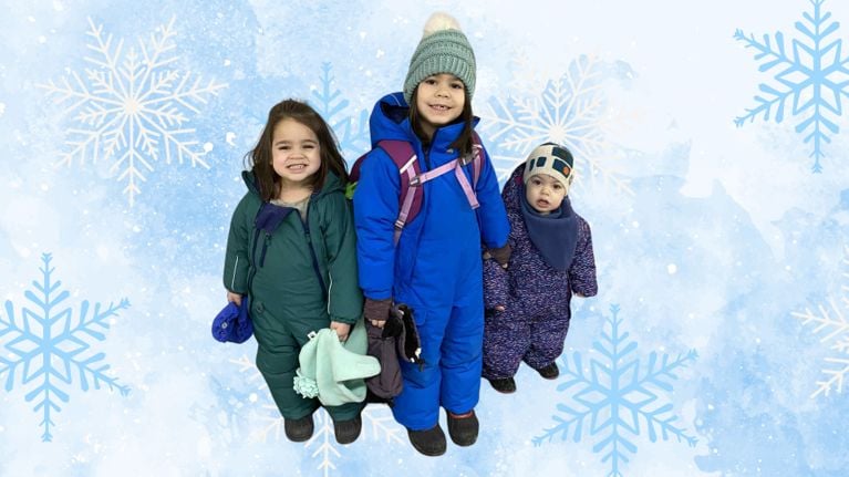 Three young children dressed in green, blue, and purple MEC Toaster Suits pose together. Two of them wear backpacks and winter hats, while the youngest looks curious and cozy.
