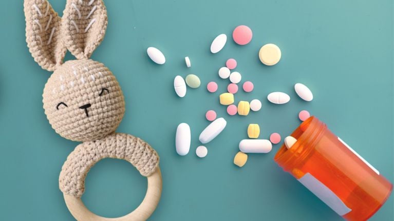 Colorful pills spill from an orange prescription bottle next to a crocheted bunny baby rattle on a teal background.