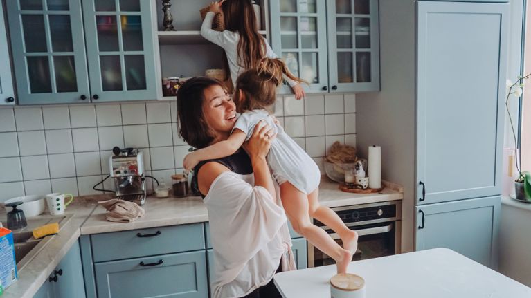 Mother smiling and holding her toddler in a cozy kitchen while another child climbs the counter to reach a cupboard.