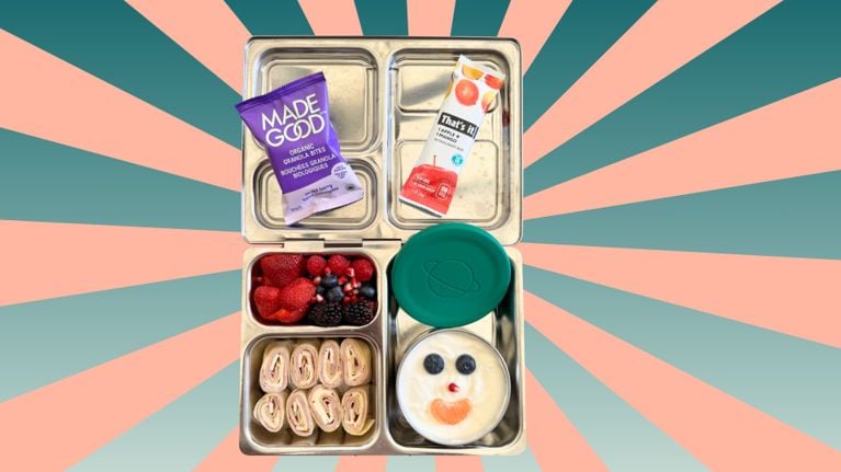 Animated GIF of a metal lunchbox being packed with turkey and cheese pinwheels, fresh berries, a yogurt cup decorated with a smiley face made of fruit, and two snack bars. The background features a teal polka dot pattern on a light green backdrop.