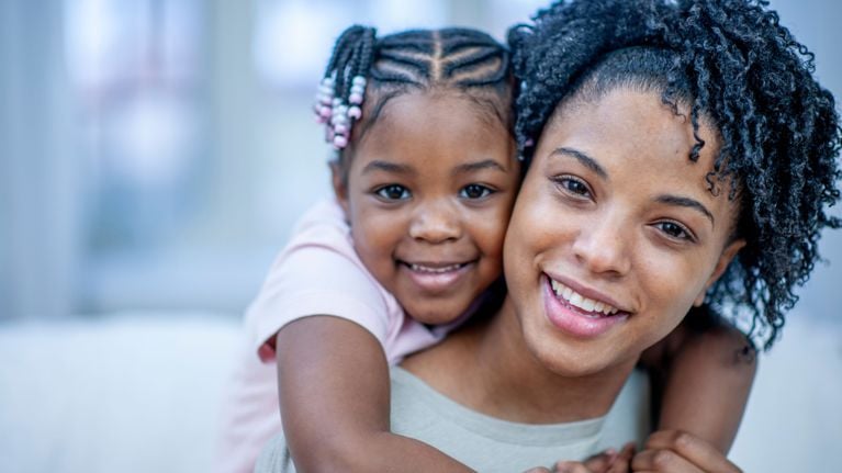 A smiling young Black girl with beaded braids hugs her mother from behind. Both are looking at the camera, close together, with joyful expressions.