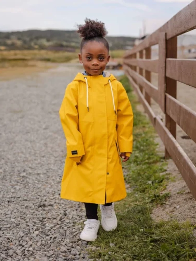 Young child standing outdoors on a gravel path beside a wooden fence, wearing a bright yellow raincoat, black leggings, and white sneakers. The background features a scenic landscape with grassy hills.