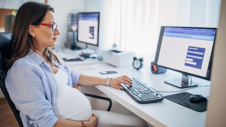 A pregnant office worker wearing glasses sits at her desk, typing on a computer and smiling slightly in a bright, modern workspace.