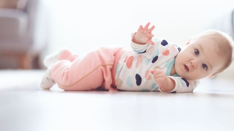 A baby in pink pants and a patterned shirt lying on their side on the floor, appearing mid-roll and looking at the camera.