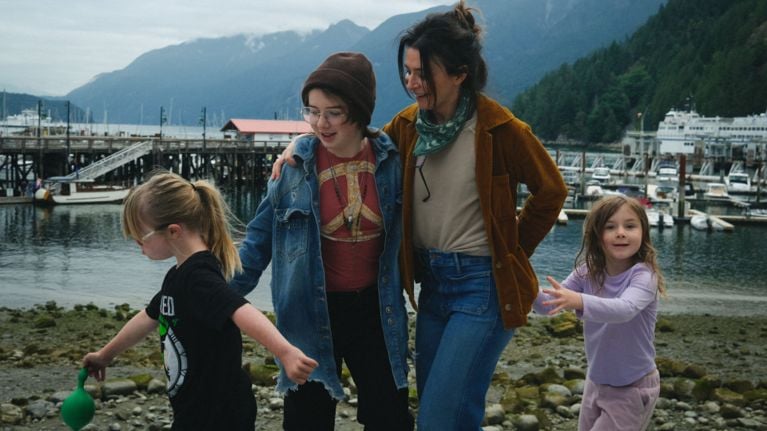 Caterina Scorsone walks along a rocky shoreline with her three daughters, smiling and holding one close as the others play. Boats and mountains fill the background on a cloudy day.