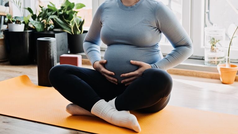 A pregnant person sitting cross-legged on an orange yoga mat, holding their belly with both hands during a prenatal exercise session at home.
