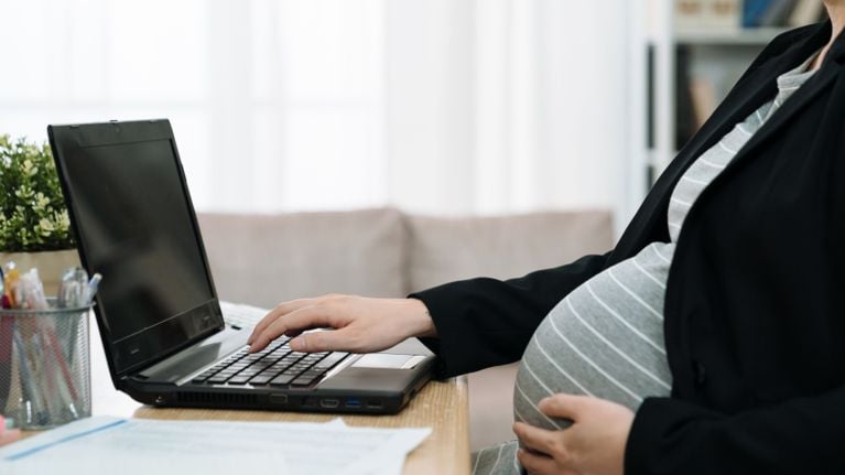 A pregnant person wearing a blazer works on a laptop at home, one hand on their belly and the other on the keyboard.