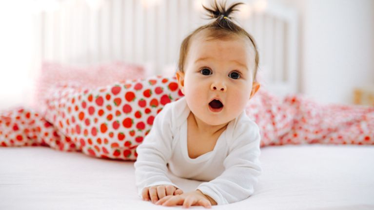 A baby with a small ponytail lying on their stomach on a bed, looking wide-eyed with an open mouth.