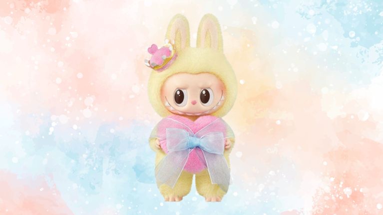 A yellow plush Labubu figure wearing a tiny crown with a heart and holding a large pink heart-shaped cushion wrapped with a blue ribbon bow. The background is pastel watercolor in peach, blue, and cream tones.