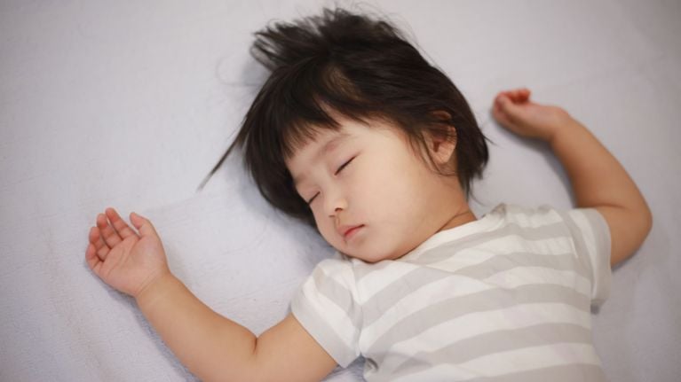 A toddler with dark hair sleeps on their back, arms spread out, wearing a white and gray striped shirt on a light-colored bed.