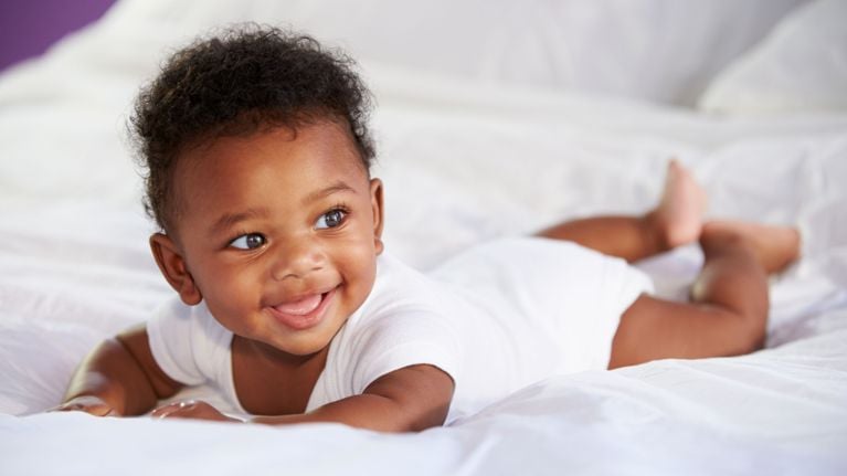A baby in a white onesie lying on their tummy on a bed, smiling brightly and lifting their head.