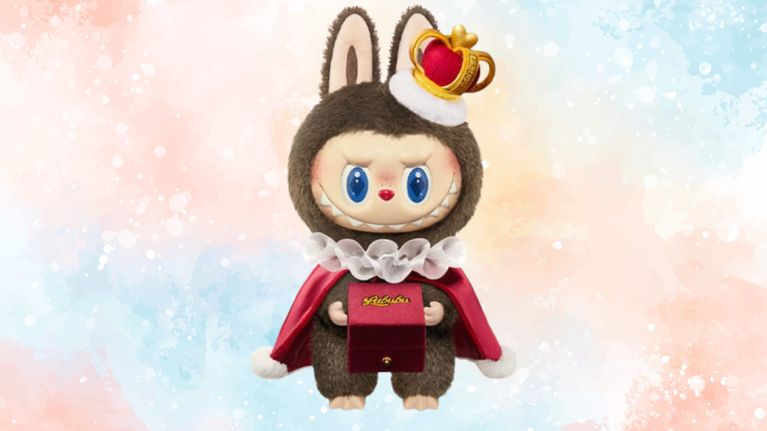 A brown Labubu plush dressed in a regal outfit with a red cape, white ruffled collar, and a crown. The doll holds a red box labeled “Labubu.” The background is soft pastel watercolor.