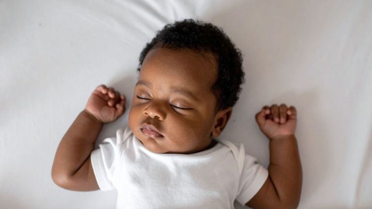 A baby sleeps on their back in a white onesie, with both fists gently clenched and resting near their head.