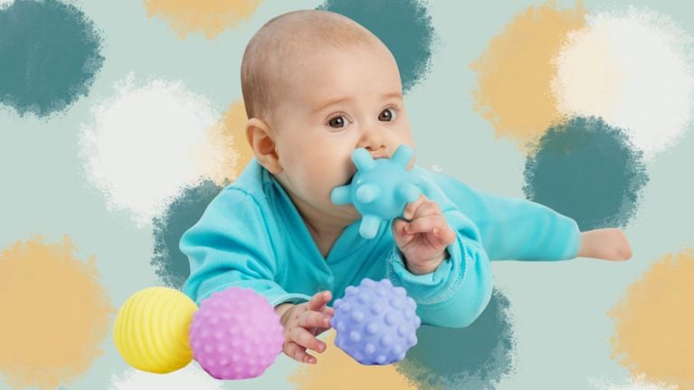 Baby in a teal outfit lying on stomach, chewing on a blue textured teether with other sensory balls nearby.