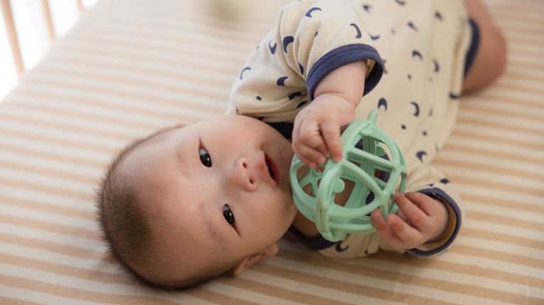 A baby lying on their side on a striped mattress, holding a green toy ball and looking up.