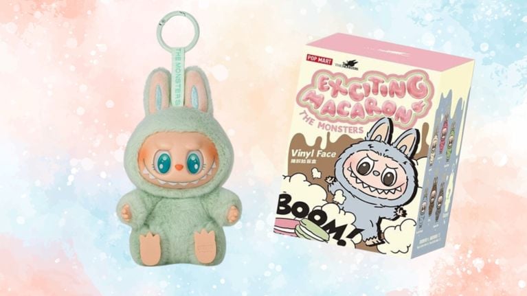 A small green Labubu plush keychain with big teal eyes and rabbit ears, shown beside its Pop Mart “Exciting Macaron” blind box packaging. The background is pastel watercolor in soft pink and blue hues.