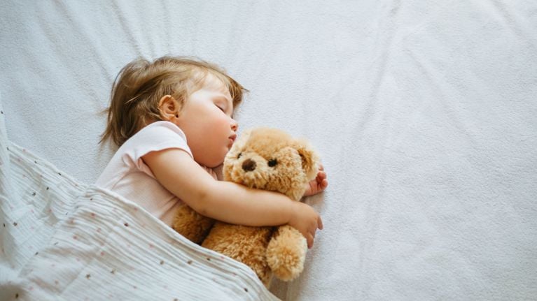 A toddler sleeps on their side, cuddling a soft brown teddy bear under a white blanket with small star patterns.