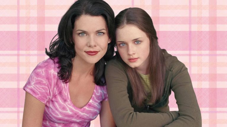 Lorelai and Rory Gilmore, the mother and teenage daughter from the TV show Gilmore Girls, sit close together against a pink plaid background. Lorelai, with dark hair and a pink tie-dye top, smiles warmly beside Rory, who has long brown hair and wears a green hoodie. The image captures their close, affectionate mother-daughter bond.