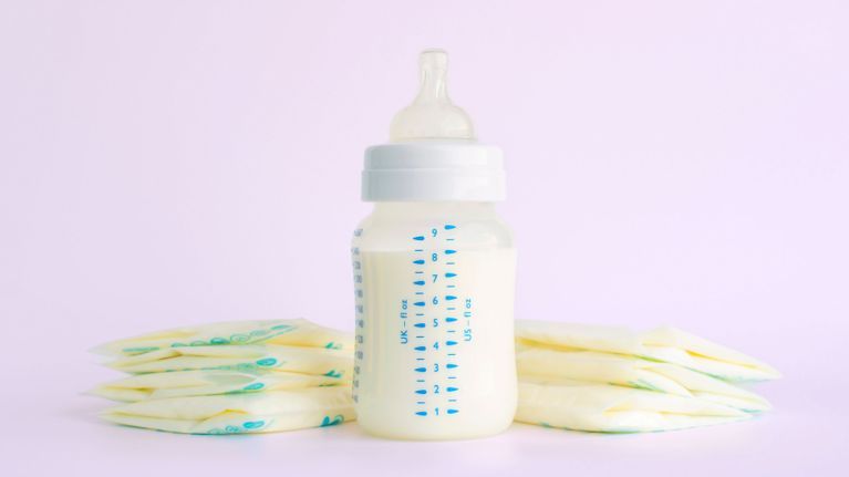 A baby bottle filled with breast milk sits on a pink background, surrounded by several frozen breast milk storage bags.
