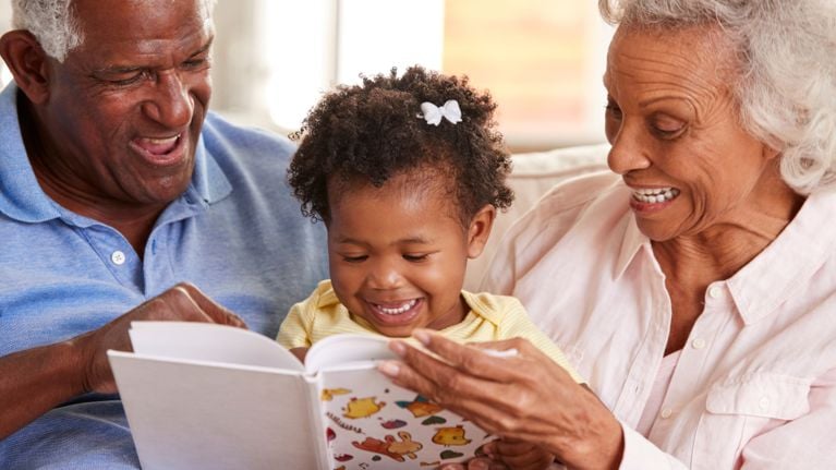 Grandparents smiling and reading a picture book with their young grandchild, sharing a joyful moment together on the couch.