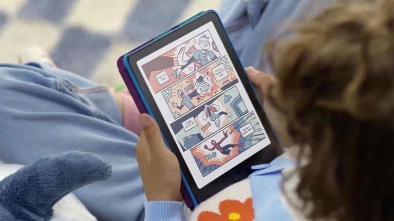 A child reclines on a couch reading a colourful comic on the new Kindle Kids with a colour E Ink display. The page shows a vibrant, cartoon-style superhero story