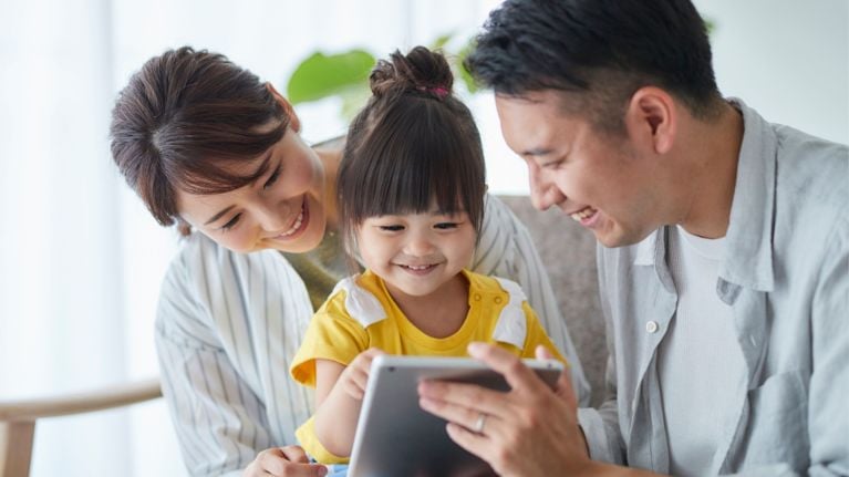 Parents and their young child smiling while using a tablet together at home, enjoying screen time as a family activity.
