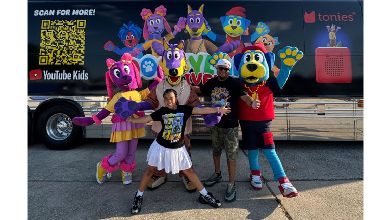 Claude Brooks and his daughter pose with three life-size Doggyland character mascots in front of a large tour bus decorated with the show’s artwork and a YouTube Kids logo.