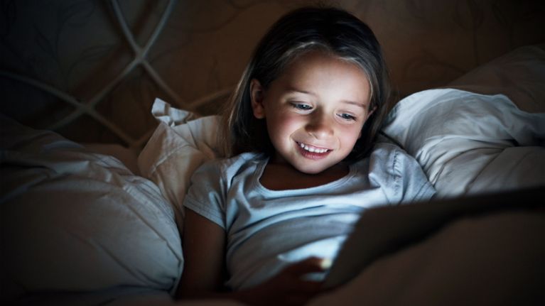 Child lying in bed at night, smiling while watching something on a tablet that glows softly in the dark.