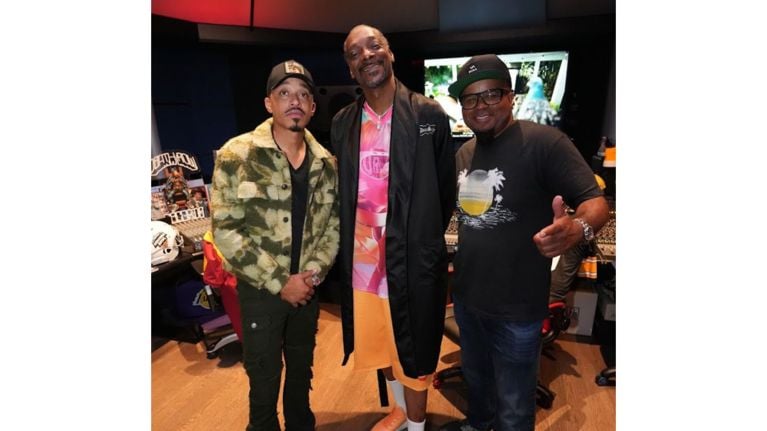 Three adults, including Snoop Dogg, stand together smiling inside a music studio filled with equipment and posters.