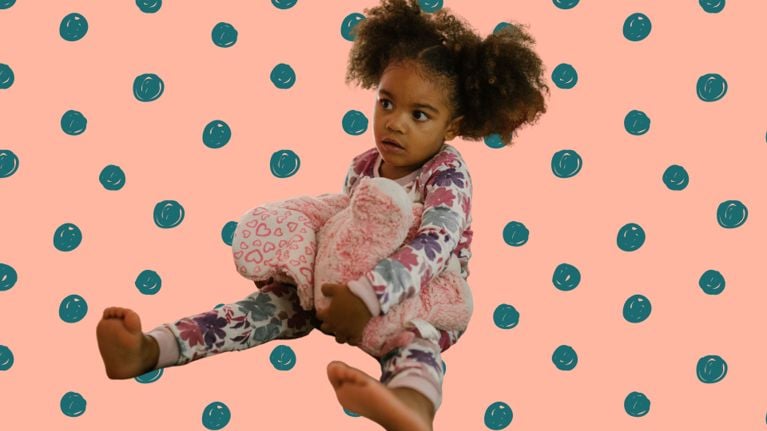 A young child in patterned pajamas holds a pink stuffed animal while sitting against a peach background with teal polka dots, looking slightly off to the side.