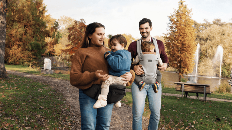 A mother and father wearing baby carriers walk outside during the fall.