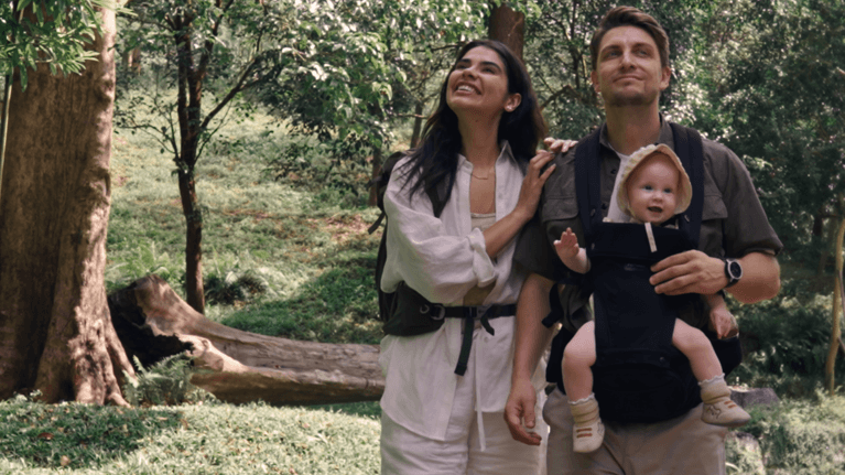 Young parents walk in a forest with the father wearing a baby carrier. The baby is smiling and the parents admire the nature around them.