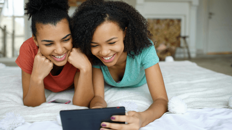 Two female teens look at their tablet while laying on a bed.