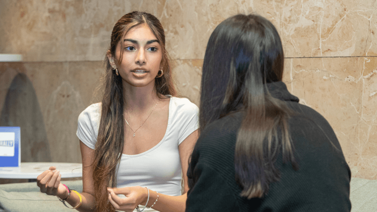 A young woman wearing a white shirt speaks to another young woman wearing a black jacket.
