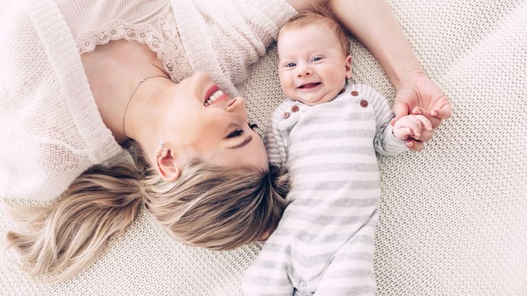 Mother lying on a soft blanket, smiling while holding hands with her happy baby dressed in striped pajamas, capturing a warm and tender bonding moment.