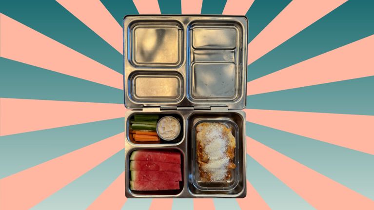 A stainless steel bento-style lunchbox filled with slices of watermelon, carrot and cucumber sticks with a small cup of dip, and a portion of cheesy lasagna sprinkled with grated parmesan. The lunchbox sits on a background of teal and coral sunburst stripes.