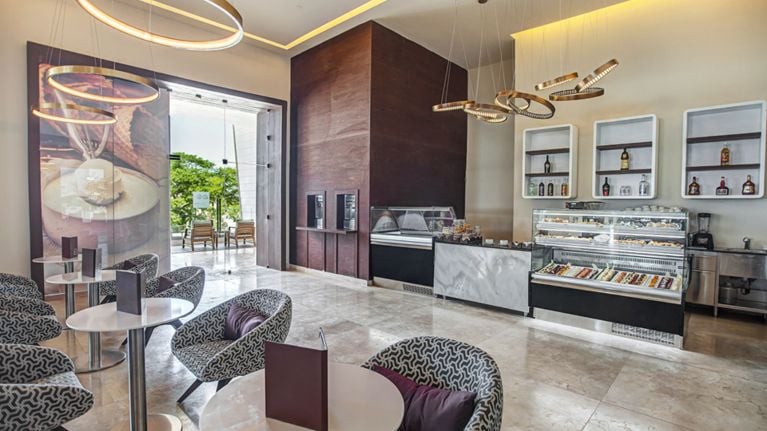 A sleek café with marble floors, patterned lounge chairs and small round tables. A pastry display case sits beneath shelves of bottles, with soft-serve machines and a large photo mural visible near the entrance to an outdoor patio.