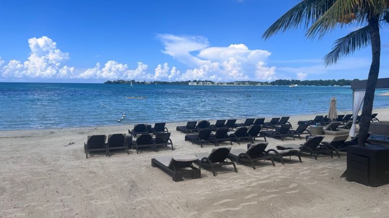 Rows of empty beach loungers sit on soft, light sand overlooking calm turquoise water. Floating buoys mark a swimming area, while palm trees frame the scene under a bright blue sky with large, billowing clouds.